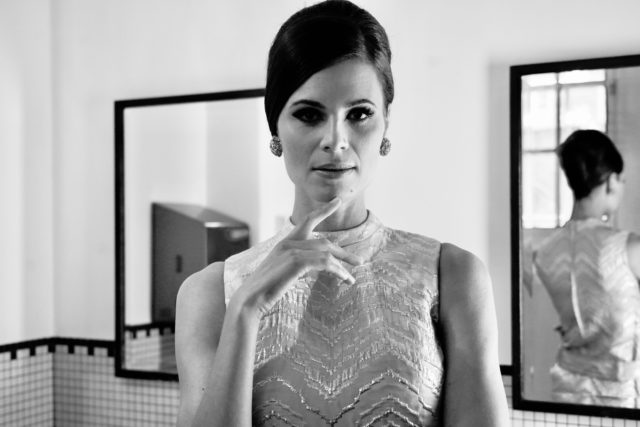 Britta Soll, Eesti Draamateatri näitleja. Mustvalge portreefoto 1960ndate stiilis riietuse ja meigiga naisest. Naine kannab sädeleva laienlise mustriga, ilma varrukateta heledat kleiti ja suuri ümmargusi kõrvarõngaid. Juuksed on hoolikalt viimistletud soengus. Naine vaatab enesekindlalt otse kaamerasse, sõrmed elegantselt lõua juures. Ta seisab teatri tualettruumis kahe suurema peegli vahel. Tema selg peegeldub taustal olevast ühest peeglist, lisades kompositsioonile sügavust.