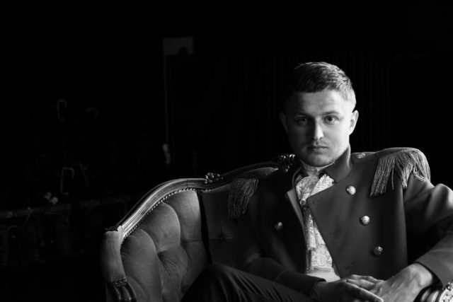 Christopher Rajaveer, Eesti Draamateatri näitleja. Mustvalge foto noorest mehest, kes istub elegantse sametkattega antiiktugitoolis. Tal on seljas teatraalne kõrgema sõjaväelase mundrit meenutav pintsak sädelevate nööpide ja narmastega õlgadel, mille all paistab pitsiline krae. Mees vaatab otse kaamerasse enesekindla ja rahuliku pilguga. Taust on tume.