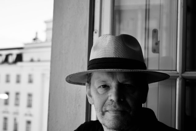 Hendrik Toompere, Eesti Draamateatri lavastaja. Pildil on mustvalge portree mehest, kes kannab heledat kaabut tumeda paelaga. Ta istub akna kõrval, millelt peegelduvad valguse ja varju kontrastid. Mehe nägu on osaliselt varjus, kuid tema silmad ja ilme on selgelt nähtavad. Taustal on näha häguselt linnahooneid, mis loovad rahuliku atmosfääri. Foto jätab vaikse ja mõtliku mulje, rõhutades lihtsust ja ajahetke intiimsust.