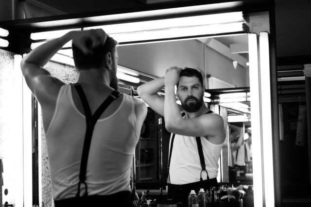 Jüri Tiiduse mustvalge portree, seisab särgi ja traksidega pükste väel peegli ees ja sätib juukseid