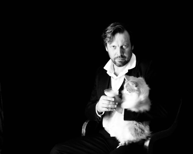 Mait Malmsten, Eesti Draamateatri näitleja. Mustvalge portreefoto mehest, kes istub käetugedega toolil. Tal on habetunud nägu, lühike tume soeng, seljas lahtise, veidi ülestõstetud kraega valge triiksärk ning must ülikond. Mees hoiab ühes käes kokteiliklaasi valge joogiga ja teises süles suurt heleda ja koheva karvaga kassi, kes vaatab klaasi poole. Mees vaatab veidi altkulmu otse kaamerasse. Fotol on tume taust, mis toob teravalt esile näo, heleda särgi, valge joogi ja heleda kassi.