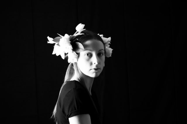 Teele Pärn, Eesti Draamateatri näitleja. Mustvalge portreefoto naisest mustal taustal. Ta seisab küljega kaamera poole, nägu otse kaamera poole. Tal on peas suur heledate õitega lillepärg. Naine vaatab otse kaamerasse rahuliku ja väljendusrikka pilguga. Tema tumedat lühikeste varrukatega riietust ja heleduse-kontrastiga valgustust rõhutab fotol tugev varjude ja valguse mäng.