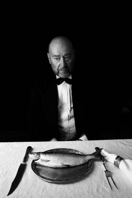 Tõnu Kark, Eesti Draamateatri näitleja. Mustvalgel fotol istub vanem mees frakis, vaadates tõsise ja keskendunud pilguga allapoole. Tema ees heleda linaga kaetud laual on elegantne taldrik, millel lebab terve kala. Laual on ka nuga, kahvel ja riidest salvrätt. Tume taust ja mehe ilme loovad pingestatud, peaaegu absurdi kalduva meeleolu.