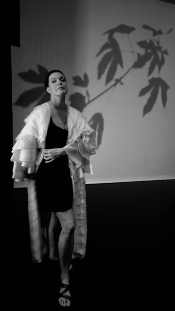 Viire Valdma, Eesti Draamateatri näitleja. Mustvalgel fotol seisab enesekindla hoiakuga naine, kes kannab lühikest tumedat kleiti ja pikka heledat, kihilise tekstuuriga hõlsti. Taustal on valgel seinal suur puuoksa ja lehtede vari.