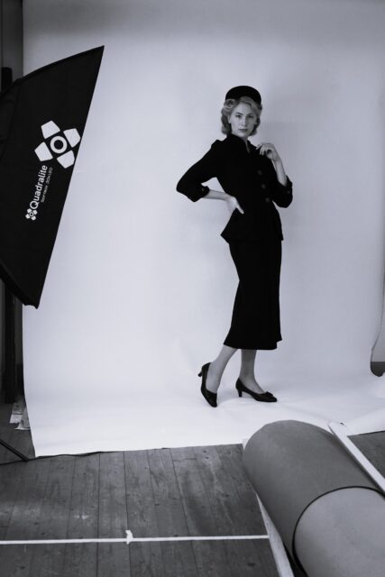 Emili Rohumaa, Eesti Draamateatri näitleja. Mustvalge foto stuudiost, kus noor naine poseerib fotostuudio valge tausta ees retrostiilis riietuses. Tal on seljas 1950ndaid meenutav elegantne kitsalõikeline kostüüm – jakk ja poolpikk seelik – ning peas väike kübar. Tal on jalas poolkõrged kontsakingad, ühte kätt hoiab puusal, teist õla juures. Vasakul on näha stuudiovalgusti ja paremas nurgas stuudiovarustust, mis rõhutavad lavatagust olustikku.