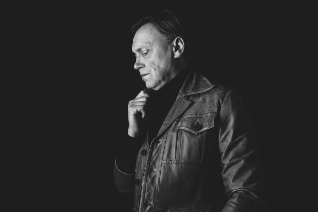 Mustvalge portreepilt Gert Raudsepast. Mustvalge portreefoto mehest, kes seisab veidi küljega kaamera poole, tema pilk on langetatud ja parema käega hoiab jaki kaelusest kinni. Tal on seljas veidi kulunud vanema moega nahktagi ja kõrge kraega tume kampsun. Valgustus toob esile tema näojooned ja rõhutab tõsist, sisekaemuslikku meeleolu.