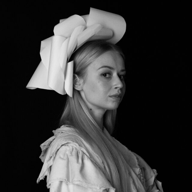 Hanna Jaanovits, Eesti Draamateatri näitleja. Mustvalgel portreefotol on noor naine, kes vaatab kergelt üle õla otse kaamerasse. Tal on pikad sirged juuksed ja peas suur skulptuurne paberist peakate. Ta kannab heledat riietust, millel on töötlemata servadega kihid. Taust on must, mis toob esile naise näojooned ja peakatte dramaatilise vormi.