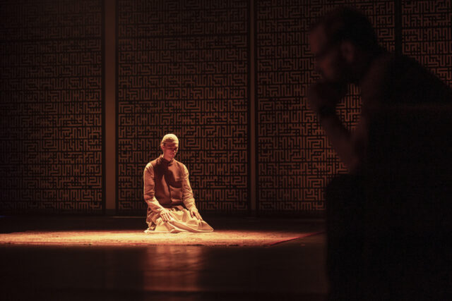 Mees põlvili palvetamas laval vaibal, taustal mustrilisest võrest sein. Lava on hämaralt valgustatud soojas oranžikas toonis. Esiplaanil paremal on hägusalt näha siluett, mis jälgib laval toimuvat. Stseen lavastusest "Ükskord Liibanonis", põlvitav mees on näitleja Richard Ester.